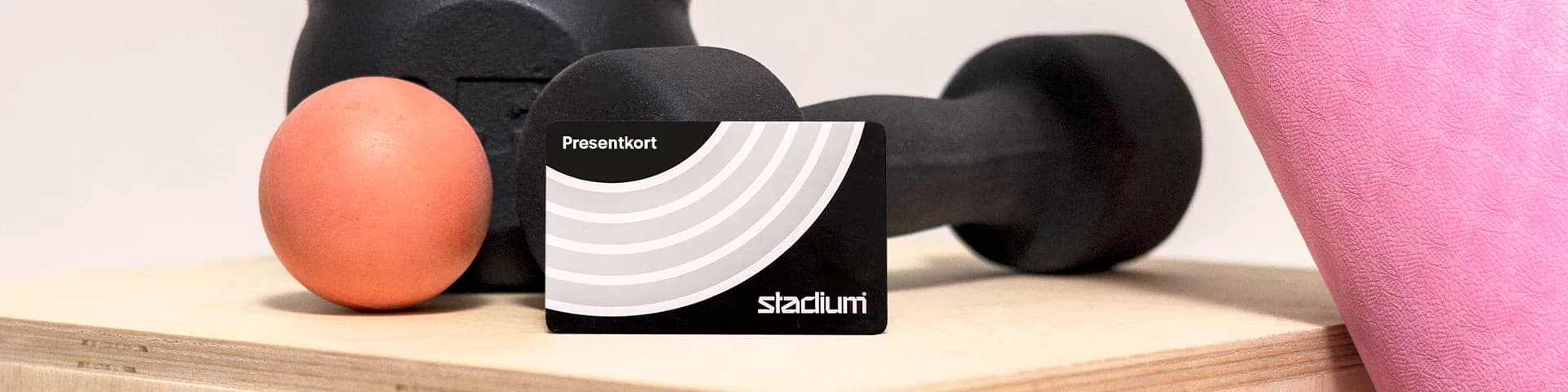 A gym equipment setup with a black dumbbell, orange ball, and pink yoga mat on a wooden surface. A gift card reads "Presentkort" and "stadium."