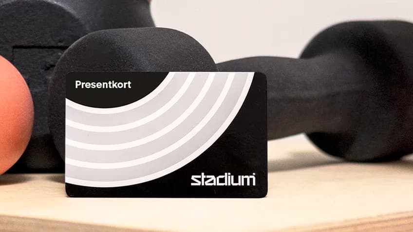 A gym equipment setup with a black dumbbell, orange ball, and pink yoga mat on a wooden surface. A gift card reads "Presentkort" and "stadium."